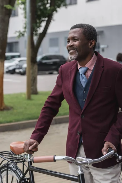 Усміхнений афроамериканський чоловік у модній бордовій куртці з велосипедом у місті — стокове фото