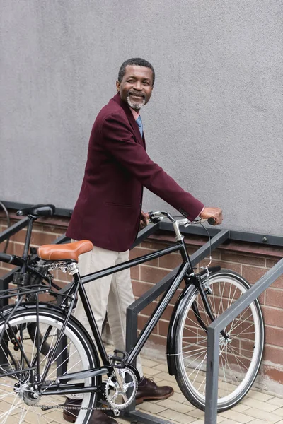 Hombre afroamericano maduro feliz con la bici en el estacionamiento para las bicicletas - foto de stock