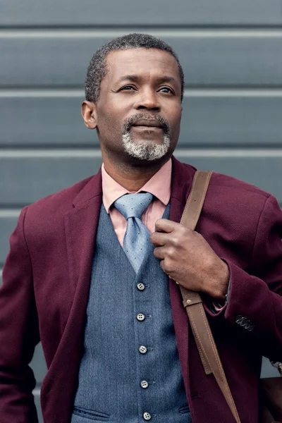 Stylish thoughtful african american man in burgundy jacket with bag — Stock Photo