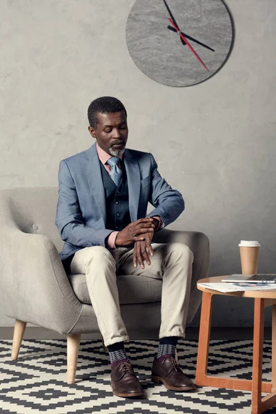 Elegante hombre de negocios afroamericano mirando reloj de pulsera mientras está sentado en la mesa con café - foto de stock