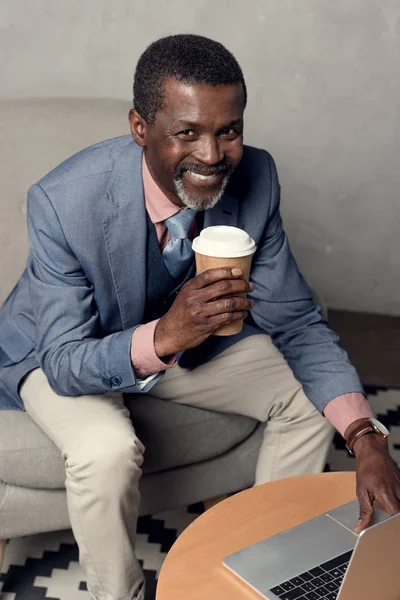 Cheerful mature african american man in blue jacket holding coffee cup and using laptop — Stock Photo