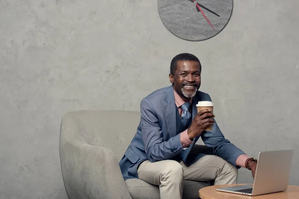 Усміхнений афроамериканський чоловік у синій куртці з кавою та ноутбуком — стокове фото