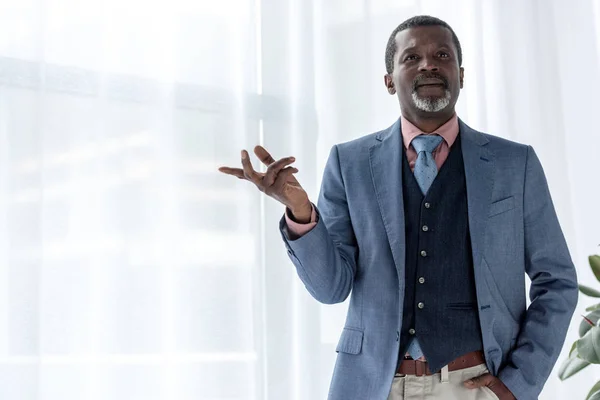 Thoughtful african american businessman in blue jacket gesturing near window — Stock Photo