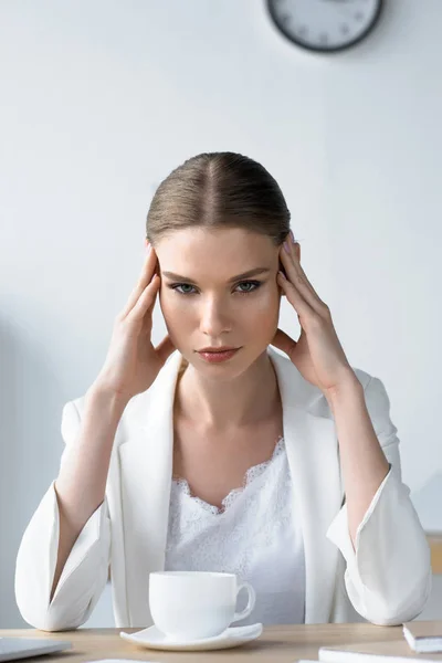 Suffering young businesswoman with headache sitting at workplace in office and holding head — Stock Photo