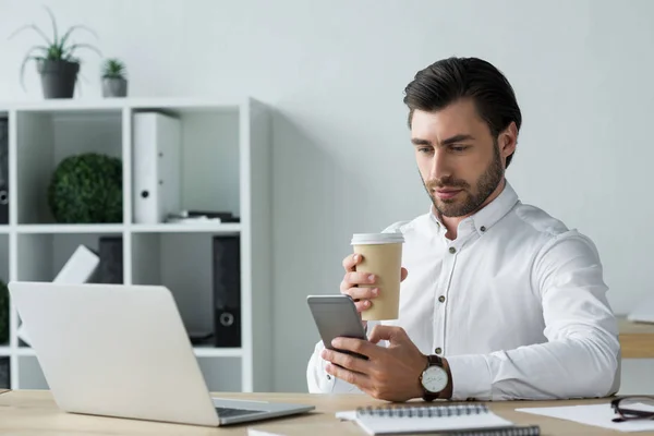 Jovem empresário bonito com xícara de papel de café usando smartphone no local de trabalho — Fotografia de Stock