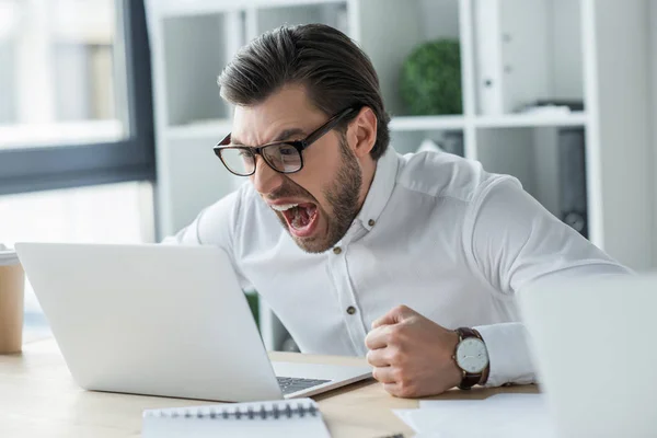 Сумасшедший молодой бизнесмен кричит на ноутбук в офисе — стоковое фото