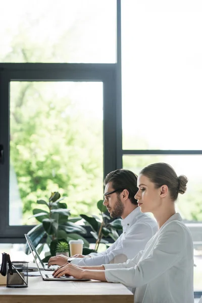 Vista lateral de los gerentes enfocados jóvenes que trabajan con computadoras portátiles juntos en la oficina - foto de stock