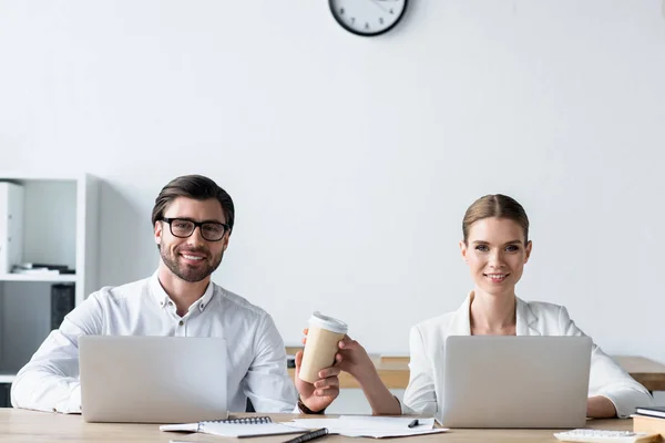 Gestionnaires travaillant avec des ordinateurs portables ensemble et passant tasse de café en papier au bureau — Photo de stock