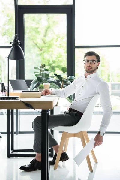 Vista lateral del joven empresario guapo sentado en el lugar de trabajo moderno en la oficina y mirando a la cámara — Stock Photo