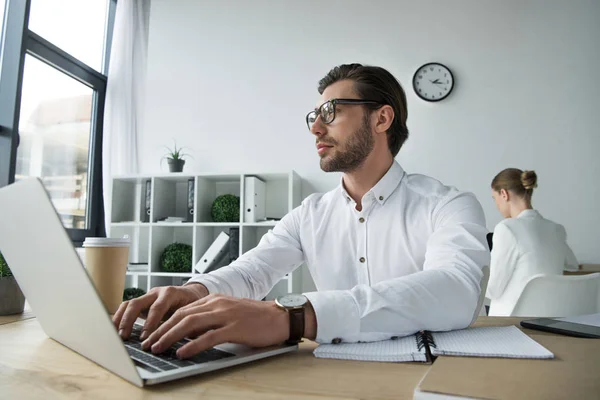 Primo piano di bel giovane uomo d'affari che lavora con computer portatile in ufficio con collega offuscato sullo sfondo — Foto stock