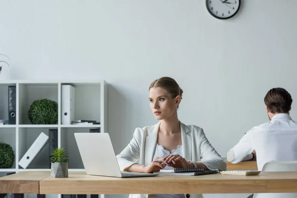 Mujer de negocios sentada en el lugar de trabajo y mirando hacia otro lado mientras su colega trabaja en segundo plano - foto de stock
