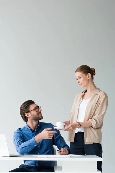 Sonriente atractiva secretaria pasando taza de café a jefe aislado en blanco - foto de stock