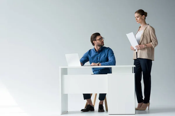 Hombre de negocios sentado en el lugar de trabajo mientras su colega de pie con documentos detrás de él en blanco - foto de stock