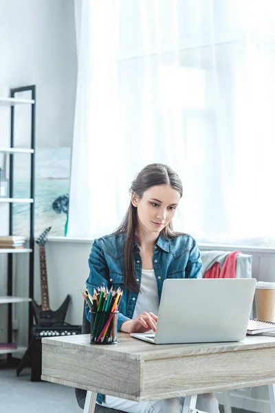 Bella ragazza utilizzando il computer portatile mentre seduto al tavolo di legno a casa — Foto stock