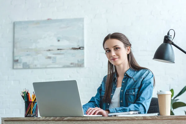 Attraktives Mädchen lächelt in die Kamera, während es Laptop am Schreibtisch benutzt — Stockfoto