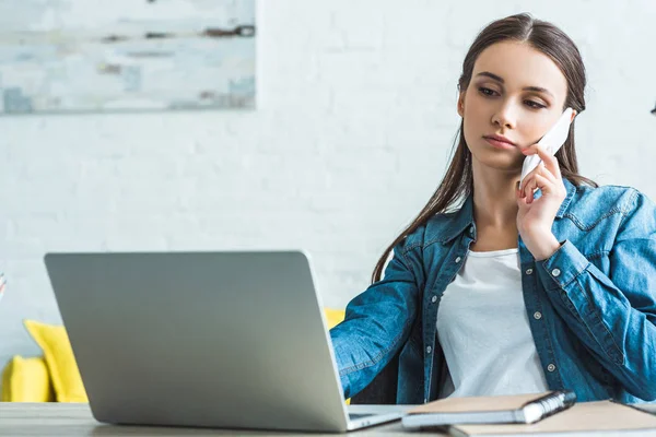 Serious teenage girl talking by smartphone and using laptop at home — Stock Photo