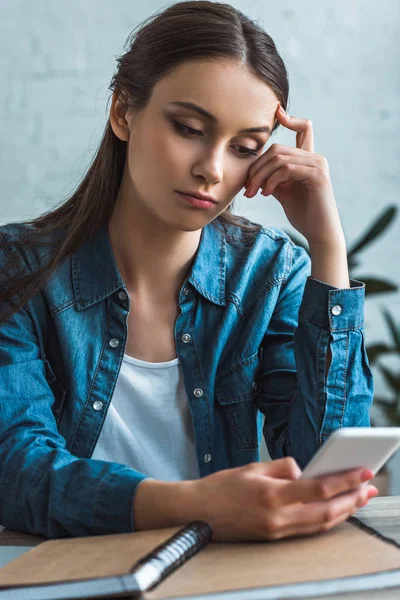 Bored teenage girl using smartphone while sitting at table with notebooks at home — Stock Photo