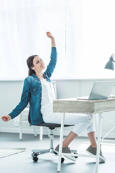 Chica feliz en los auriculares levantando la mano y mirando hacia otro lado mientras se utiliza el ordenador portátil en el escritorio - foto de stock