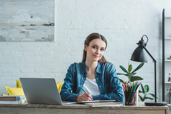 Teenage girl taking notes and smiling at camera while studying with laptop at home — Stock Photo