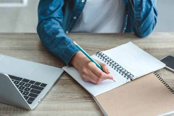 Nahaufnahme: Mädchen schreibt in Notizbuch, während sie mit Laptop am Schreibtisch lernt — Stockfoto