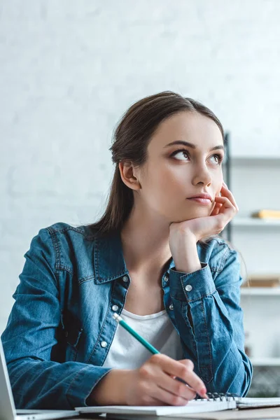 Nachdenkliches Mädchen mit der Hand am Kinn, das Notizen macht und wegschaut, während es zu Hause studiert — Stockfoto