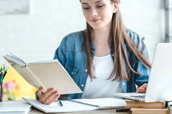 Smiling girl holding book and using laptop while studying at home — Stock Photo