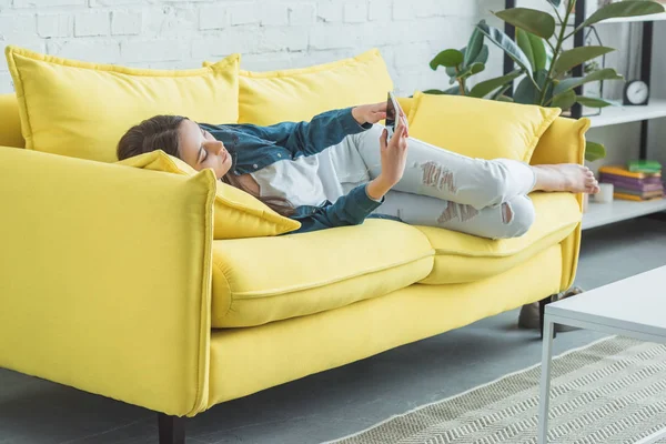 Bela adolescente deitada no sofá e usando smartphone — Fotografia de Stock