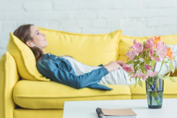 Notebook e flores em vaso na mesa e menina ouvindo música em fones de ouvido no sofá atrás — Fotografia de Stock