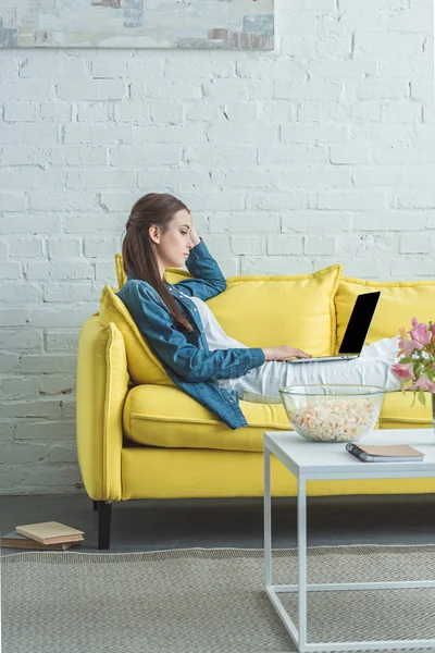 Vista lateral de la mujer joven usando el ordenador portátil con la pantalla en blanco en el sofá en casa - foto de stock