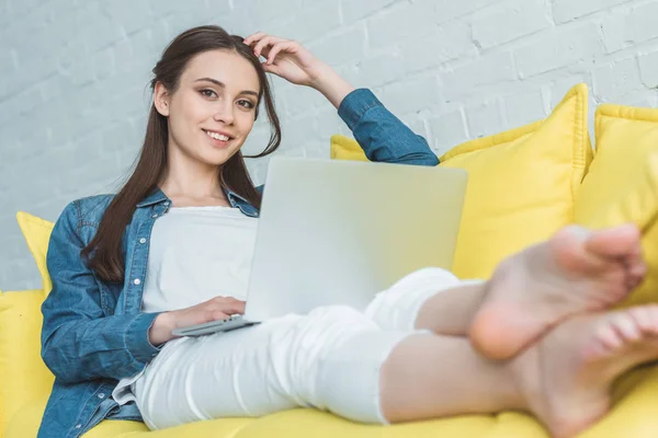Красивая молодая женщина, пользующаяся ноутбуком и улыбающаяся на камеру, сидя дома на диване — стоковое фото