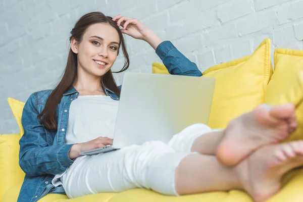 Belle jeune femme utilisant un ordinateur portable et souriant à la caméra tout en étant assis sur le canapé à la maison — Photo de stock