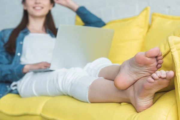 Plan recadré de sourire fille pieds nus en utilisant un ordinateur portable sur le canapé à la maison — Photo de stock
