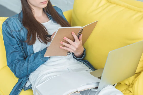 Colpo ritagliato di ragazza che scrive nel notebook e utilizza il computer portatile sul divano a casa — Foto stock