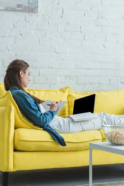 Ragazza prendere appunti nel notebook e utilizzando il computer portatile con schermo bianco sul divano a casa — Foto stock