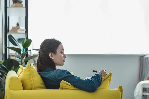 Back view of serious girl looking away while sitting on sofa and taking notes — Stock Photo