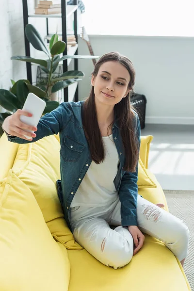 Attractive smiling girl sitting on sofa and taking selfie with smartphone — Stock Photo