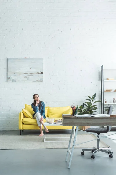 Pensive barefoot girl sitting on sofa and looking away at home — Stock Photo