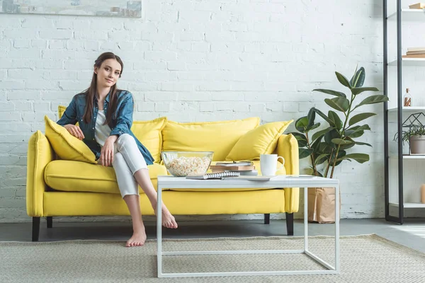 Menina descalça atraente sentado no sofá e sorrindo para a câmera em casa — Fotografia de Stock