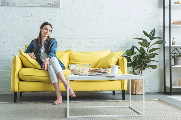 Attraktives Barfußmädchen sitzt auf dem Sofa und lächelt zu Hause in die Kamera — Stockfoto