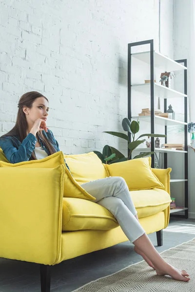Beautiful pensive barefoot girl sitting on sofa and looking away at home — Stock Photo