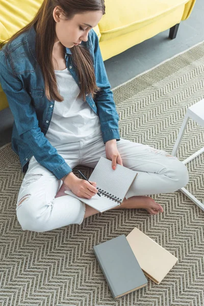 Vista de ángulo alto de la niña escribiendo en el cuaderno mientras está sentado en la alfombra y estudiando en casa - foto de stock