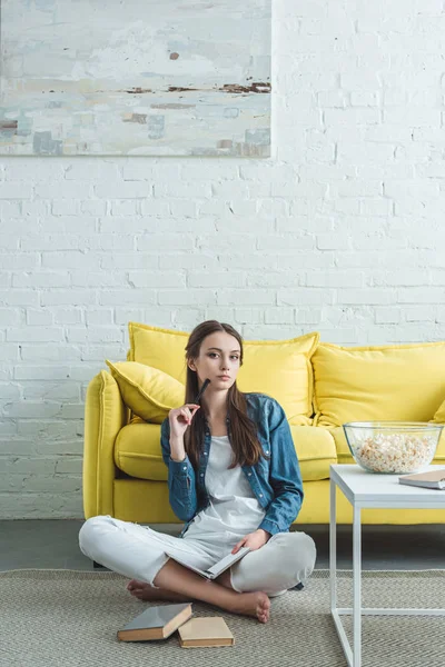 Attraktives Mädchen, das Buch in der Hand hält und in die Kamera schaut, während es auf dem Teppich sitzt und zu Hause studiert — Stockfoto