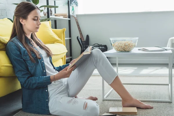 Вид збоку дівчини-підлітка читає книгу, сидячи на килимі вдома — Stock Photo