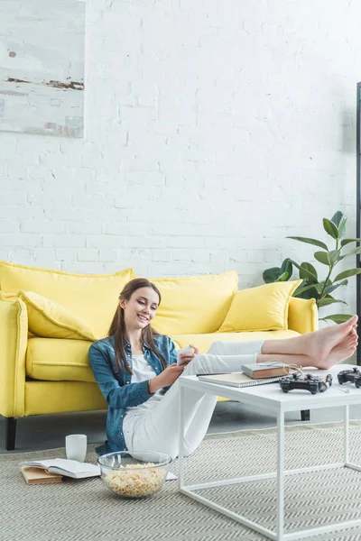 Sonriendo chica descalza sentada en la alfombra y usando el teléfono inteligente mientras estudia en casa - foto de stock