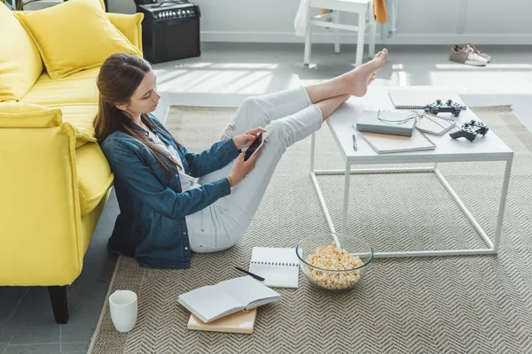 Высокий угол обзора девочки-подростка с помощью смартфона во время сидения на ковре и учебы дома — стоковое фото