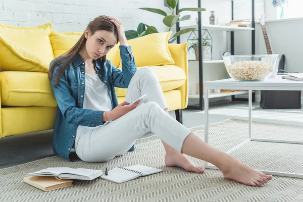 Gelangweiltes Teenager-Mädchen, das Smartphone in der Hand hält und in die Kamera schaut, während es auf dem Teppich sitzt und zu Hause studiert — Stockfoto