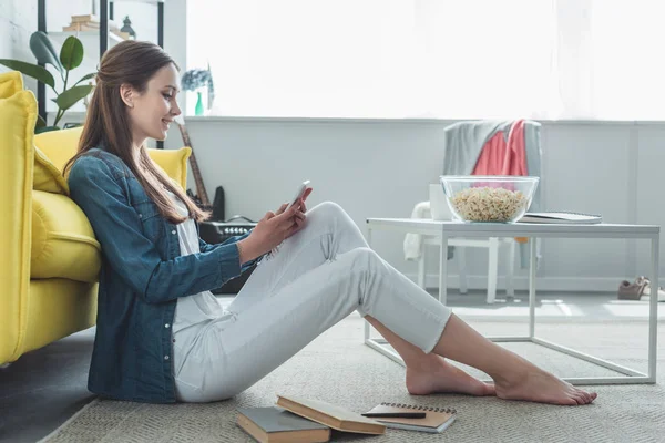 Вид збоку усміхненої дівчини-підлітка, що використовує смартфон, сидячи на килимі з книгами вдома — стокове фото