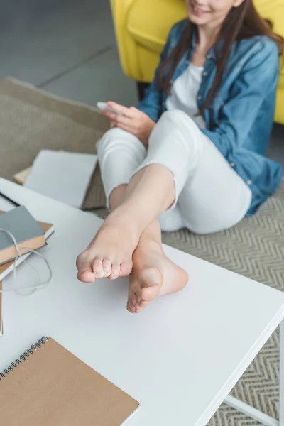 Cropped shot of smiling barefoot girl using smartphone while studying at home — Stock Photo