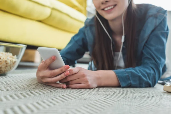 Nahaufnahme eines lächelnden Mädchens mit Kopfhörern, das sein Smartphone benutzt, während es auf Teppich liegt — Stockfoto