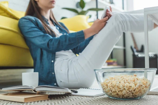 Vista de cerca de tazón de vidrio con palomitas de maíz y libros, niña usando el teléfono inteligente detrás - foto de stock